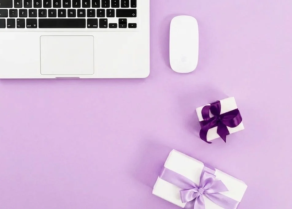 Silver laptop and white mouse on a lavender surface with two white gift boxes tied in purple ribbons
